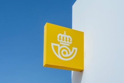 A yellow postal sign with a crown logo against a clear blue sky, representing mail services.