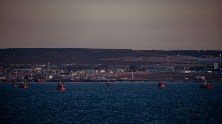 Distant view of fishing boats in a harbor at sunset with an industrial backdrop.