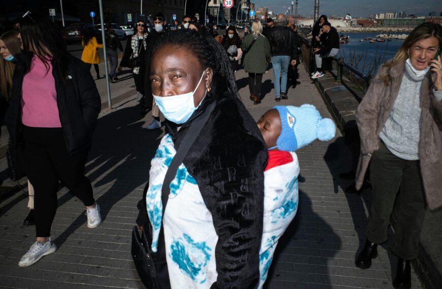 A mother carrying her child walks along a busy city sidewalk, with people wearing face masks.