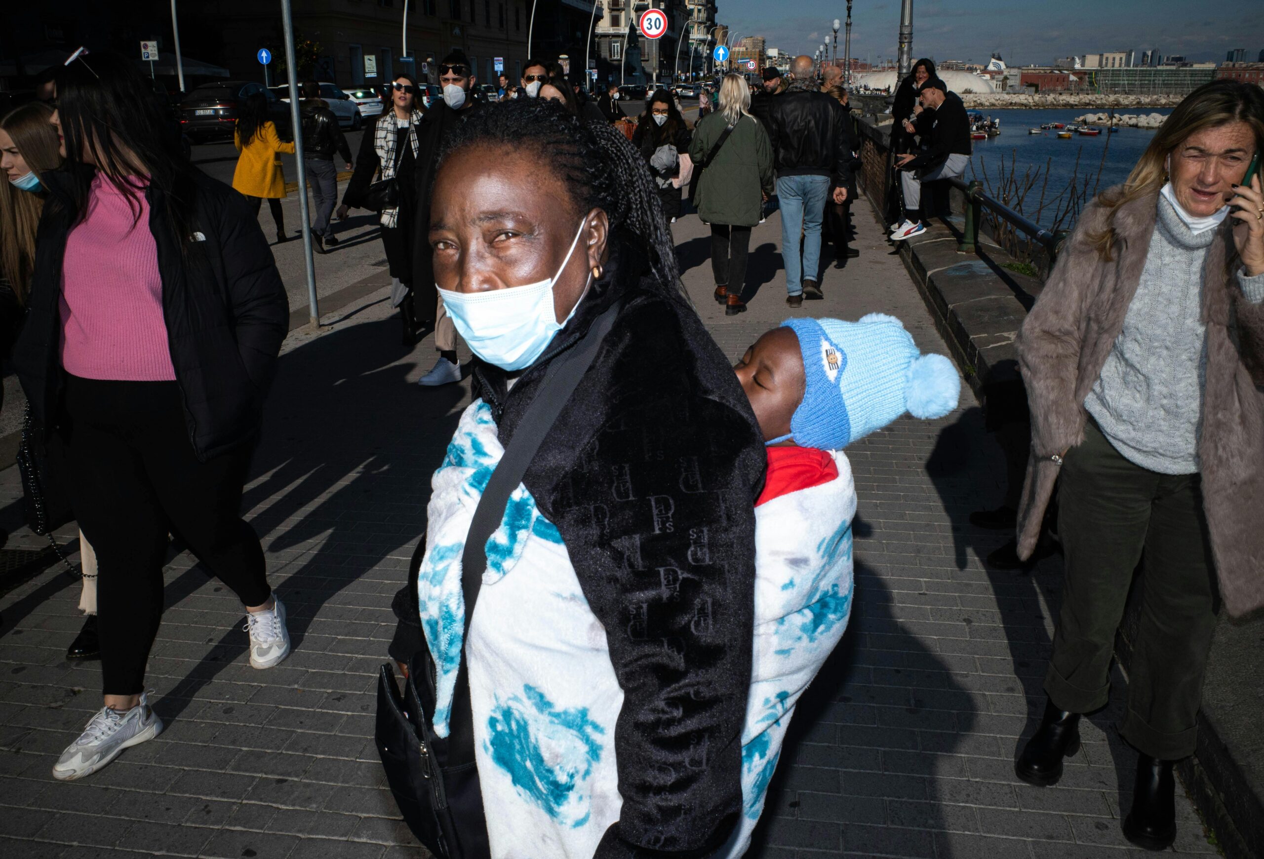 A mother carrying her child walks along a busy city sidewalk, with people wearing face masks.